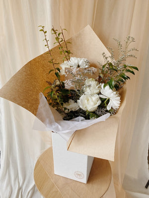 A white and green botanical flower arrangement wrapped in kraft paper displayed in an upright delivery box