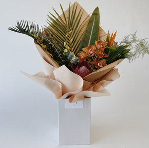 A tropical flower bouquet with palm leaves, orchids, protea and birds of paradise flower displayed in an upright delivery box and wrapped in kraft paper